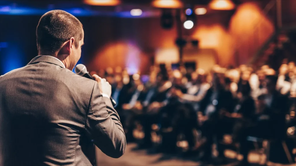 Palestrante de costas segurando microfone diante de plateia em evento corporativo