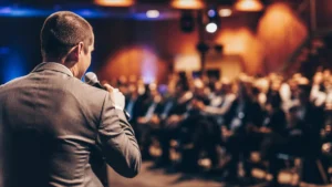 Palestrante de costas segurando microfone diante de plateia em evento corporativo